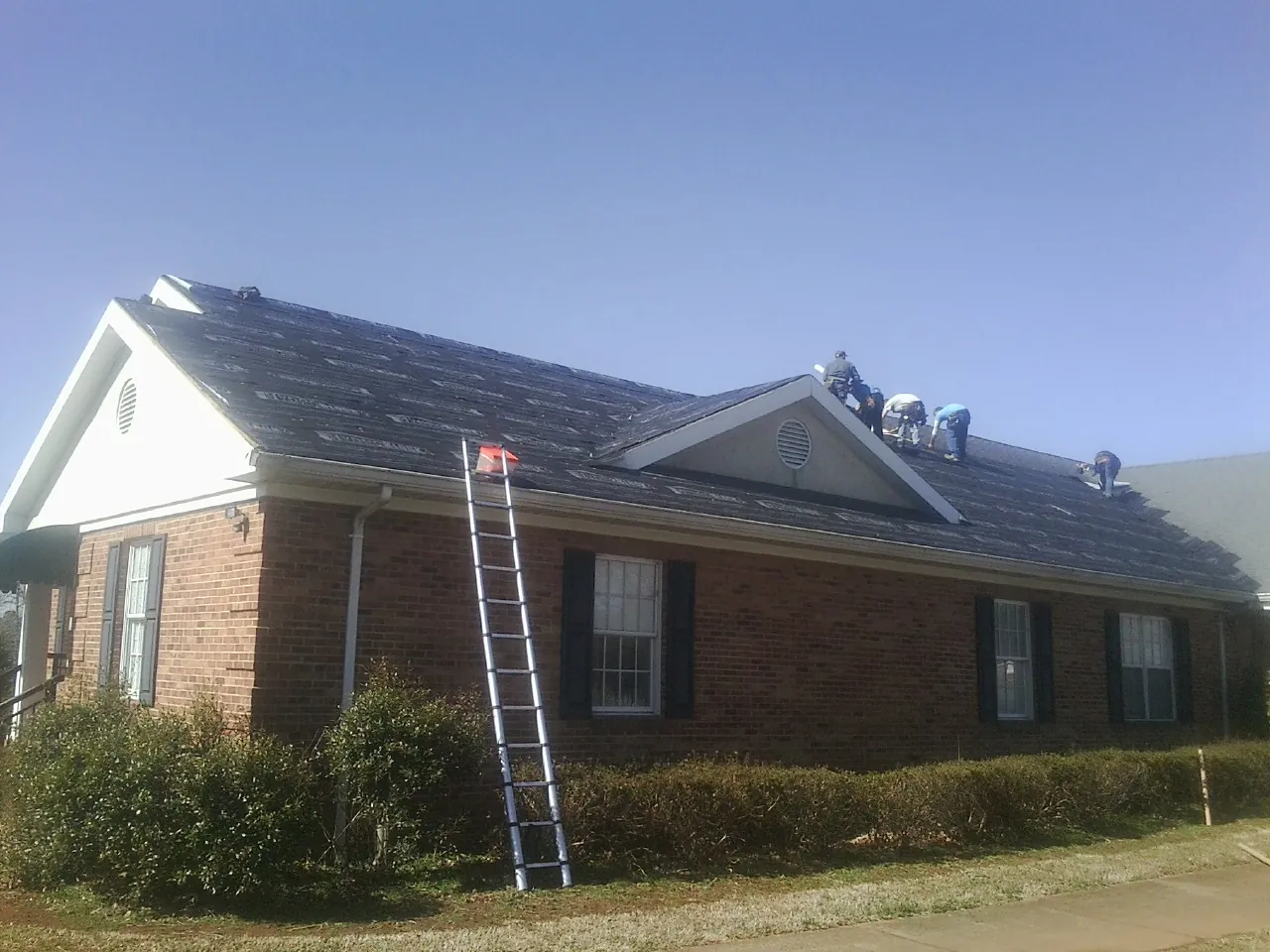 Crew tearing shingles off a brick ranch roof with a ladder set — Metal Roof Repair work by our crew in Friendswood