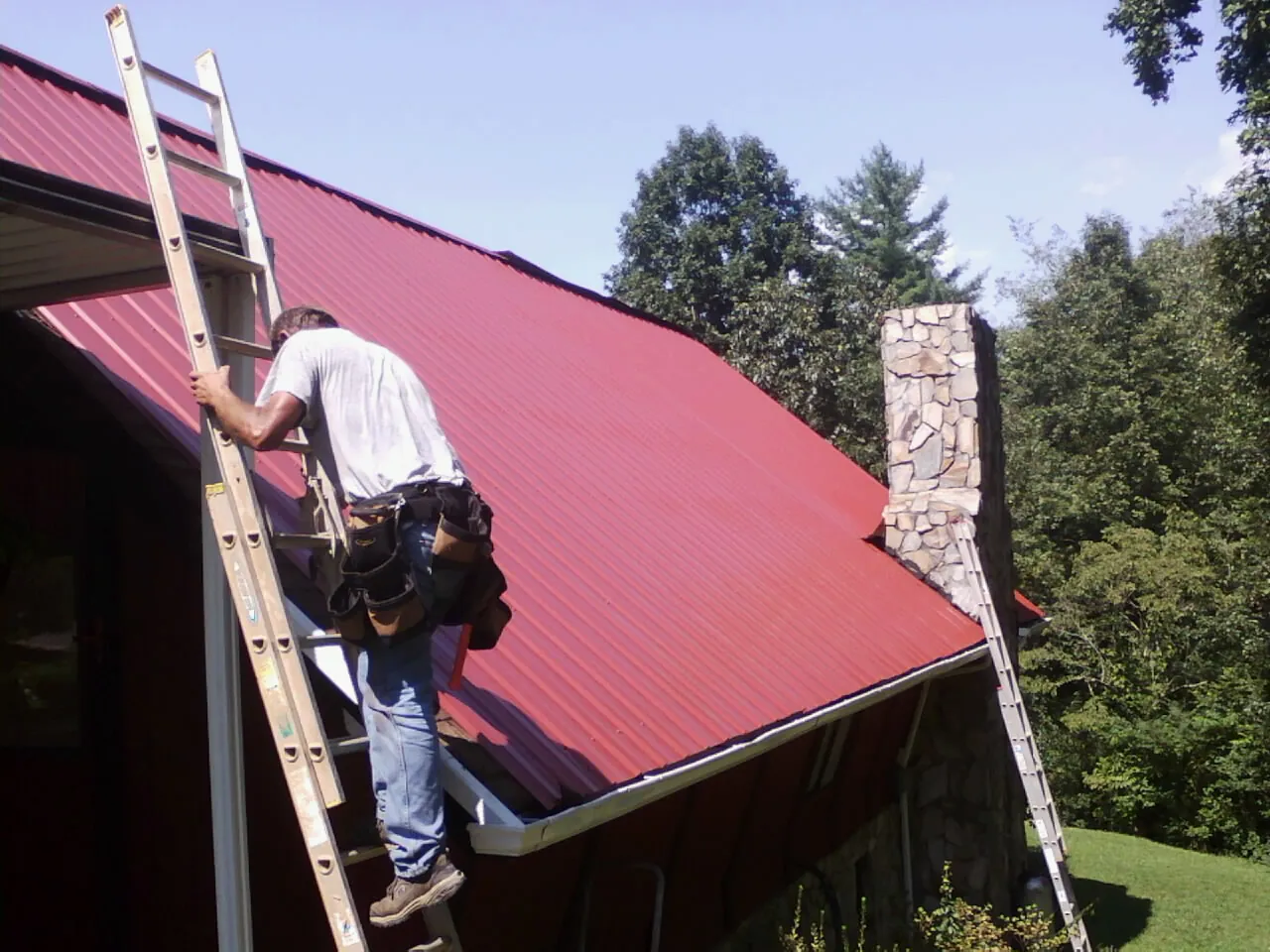 Roofer on a ladder beside a red metal roof and stone chimney — Soffit & Fascia Repair by Roofing Brothers in Friendswood